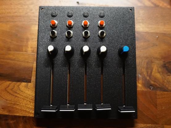 The printed black front panel on a wooden desk, with a slightly grainy, textured surface. Four columns each have an LED in a neoprene holder, a red and a black push button, a white knob and a long fader. A fifth column on the right has a blue knob and another fader.