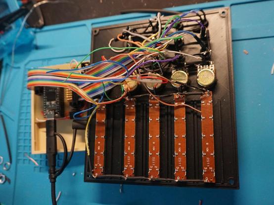 A black front panel from the backside. and a stripboard with a microcontroller next to it. The components in the panel are soldered with wires. The single wires on one end form a ribbon cable on the other end which is connected with connectors on the stripboard.
